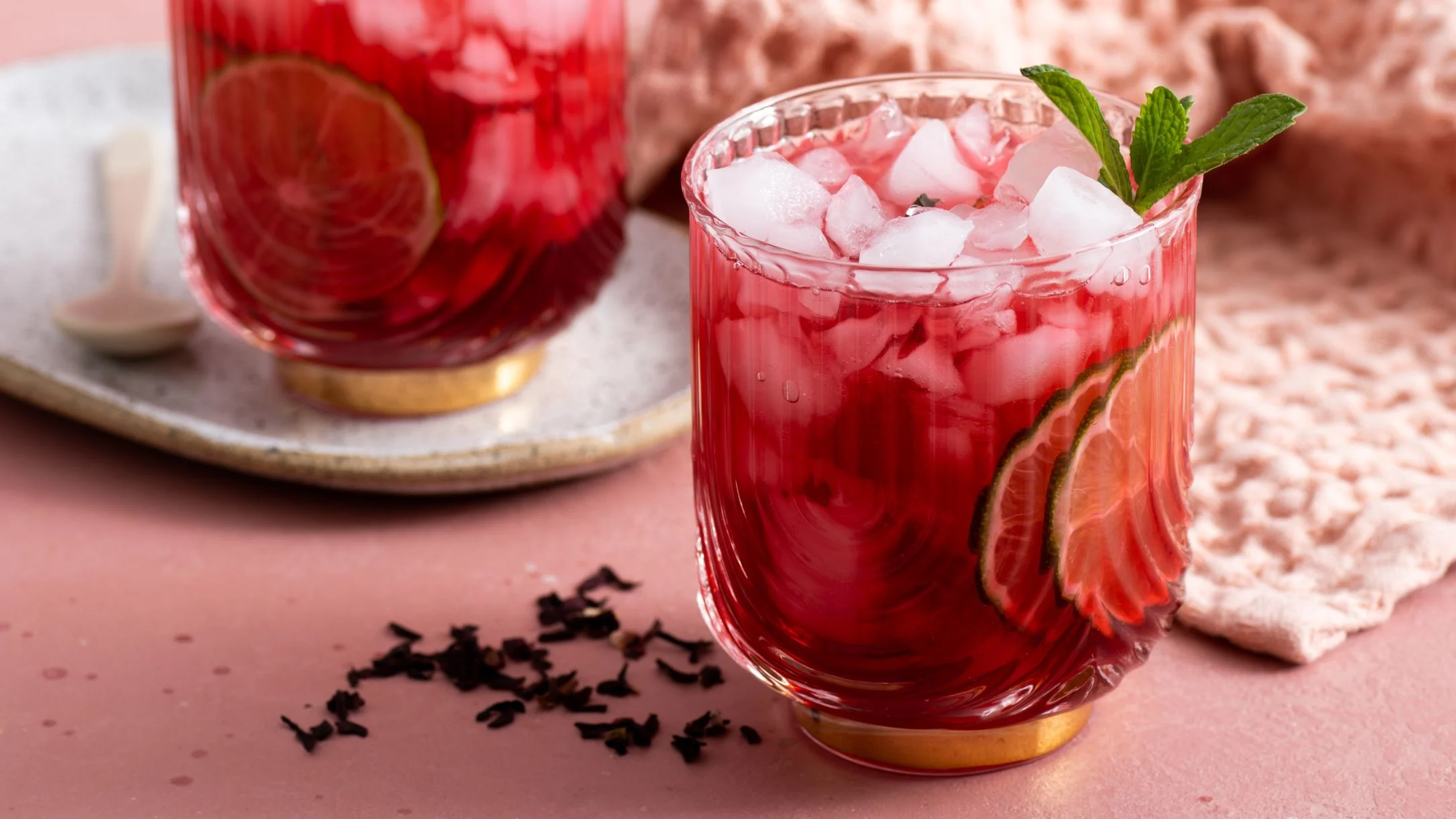 Red mocktail with ice cubes, lime slices and mint garnish in a crystal glass, with tea leaves scattered nearby.