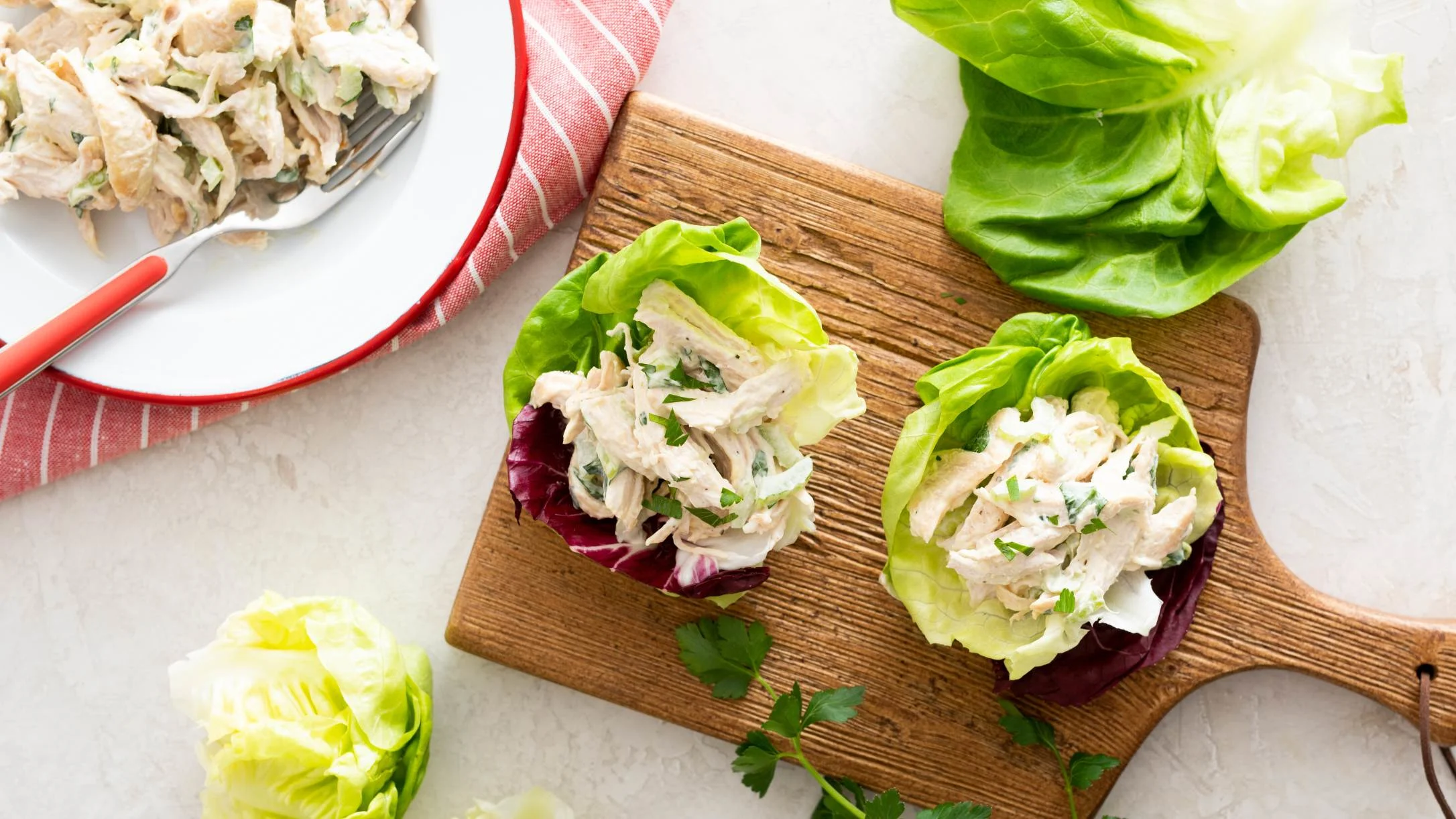 Chicken salad served in lettuce cups on a wooden board, with a bowl of additional chicken salad nearby.