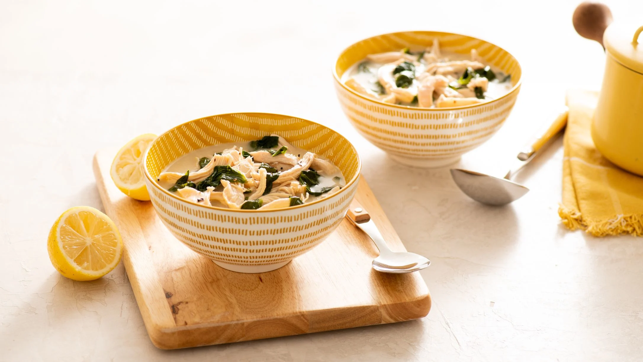 Two yellow-patterned bowls of chicken soup with greens served on wooden board with lemon slices and spoons nearby.