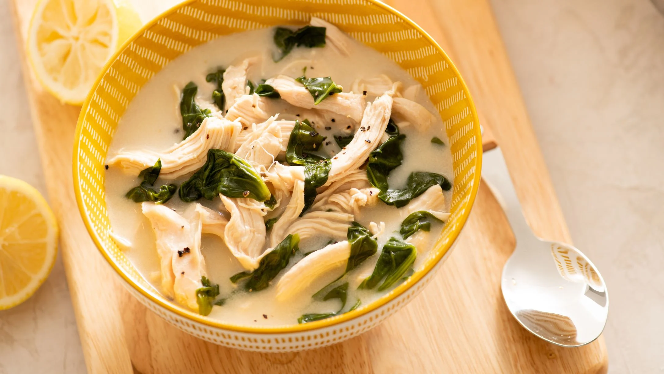 Creamy chicken soup with spinach in yellow bowl, served with lemon slices on wooden board with spoon.