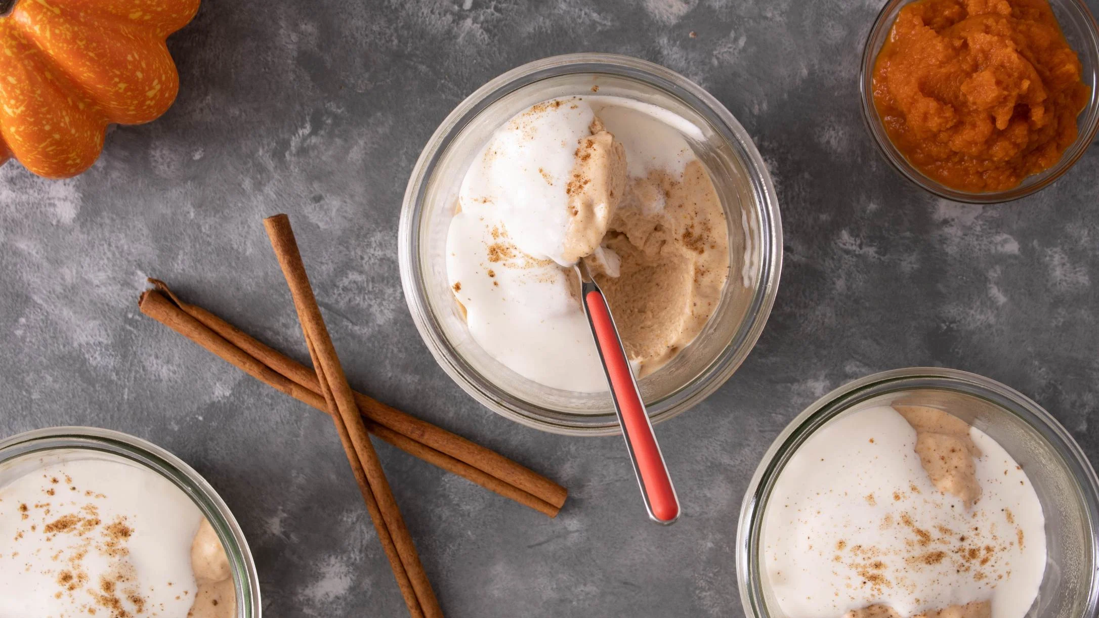 Bowls of pumpkin pudding with cinnamon sticks, a red spoon, and pumpkin puree on a gray stone surface.