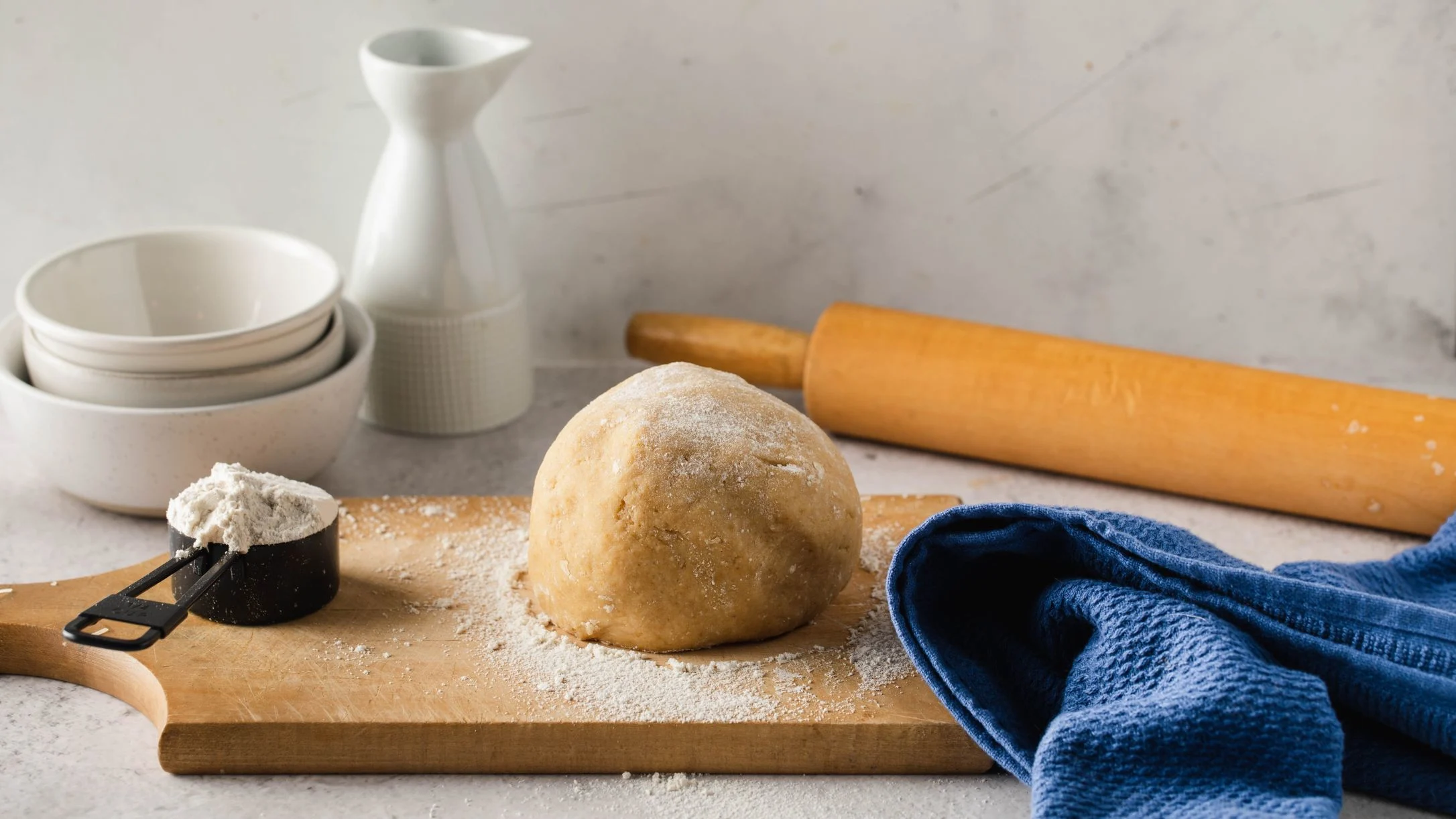 Ball of dough on wooden board with flour, rolling pin, measuring cup, white ceramic bowls, and blue kitchen towel.