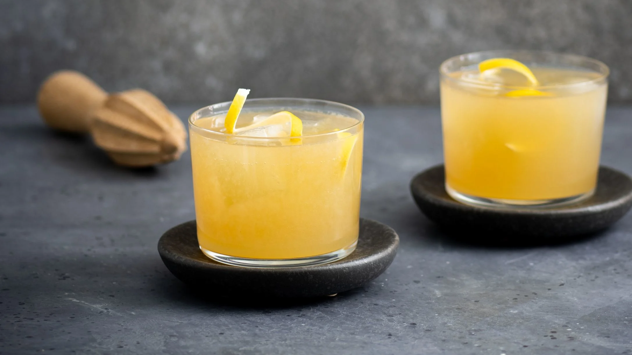 Two yellow citrus cocktails with ice and lemon garnish on dark stone coasters, wooden juicer in background.