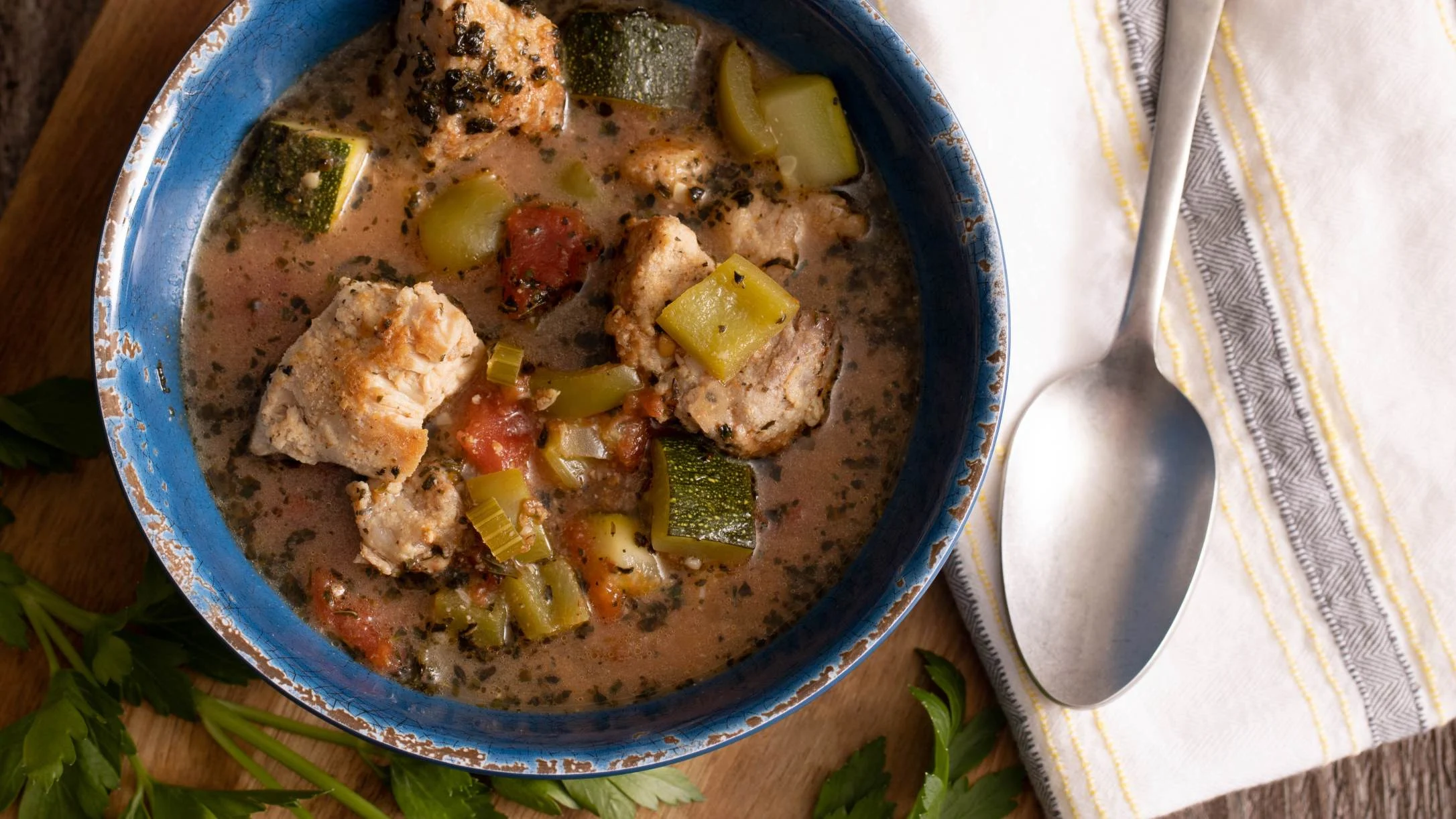 Hearty stew with chunks of meat, zucchini, and tomatoes in a creamy broth, served in a blue ceramic bowl with spoon.