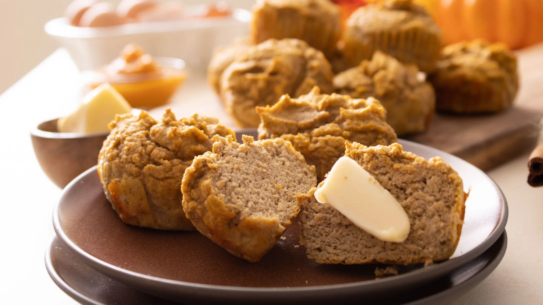 Freshly baked pumpkin muffins on a brown plate with a pat of butter melting on a sliced muffin, more muffins in background.