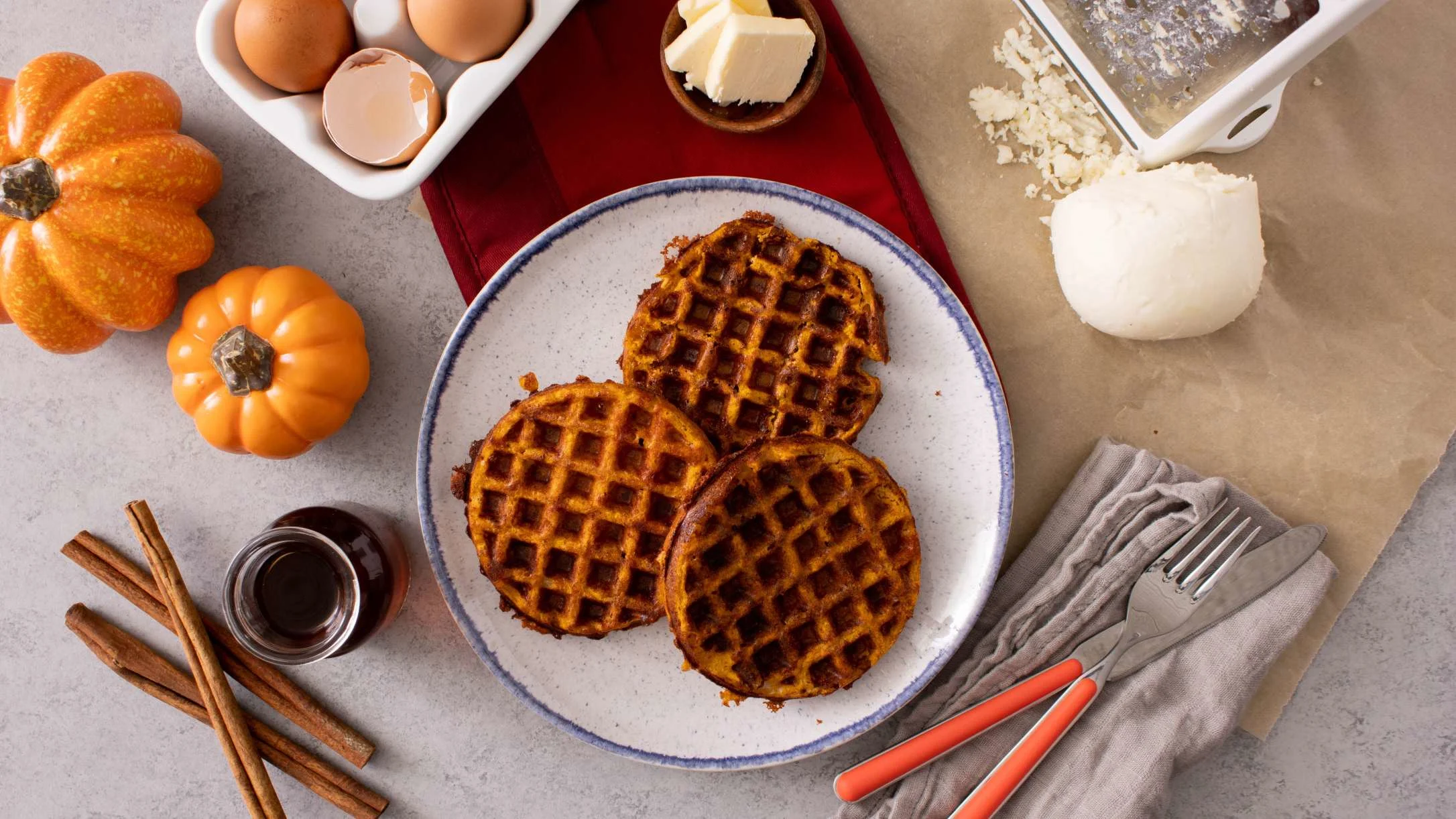 Pumpkin waffles on blue plate surrounded by baking ingredients including eggs, butter, mini pumpkins, and cinnamon sticks.