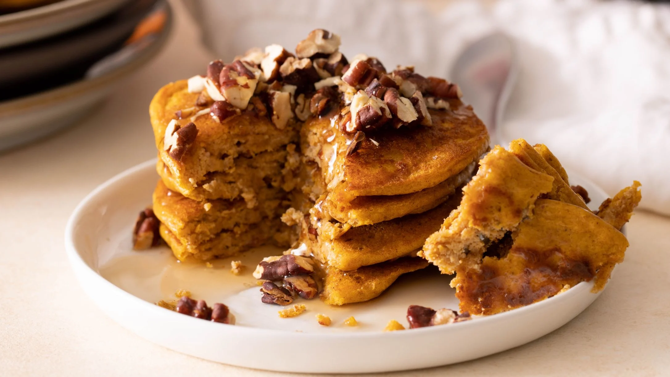 Stack of golden pumpkin pancakes with a bite taken out, topped with chopped pecans on a white plate.