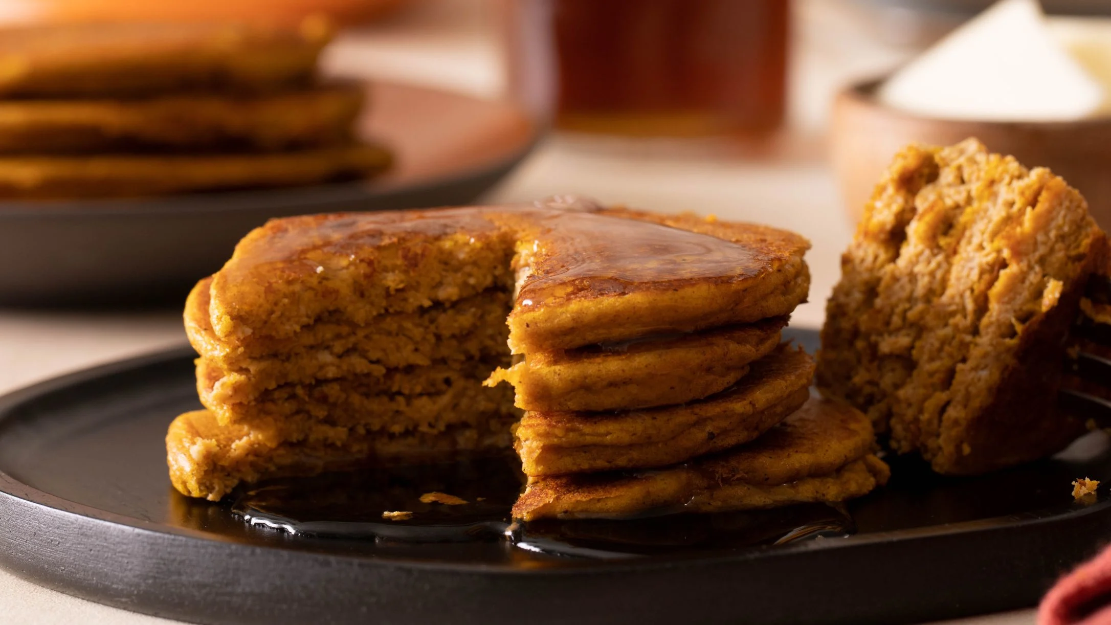 Stack of golden pumpkin pancakes on a dark plate with syrup, one pancake cut to show fluffy interior texture.