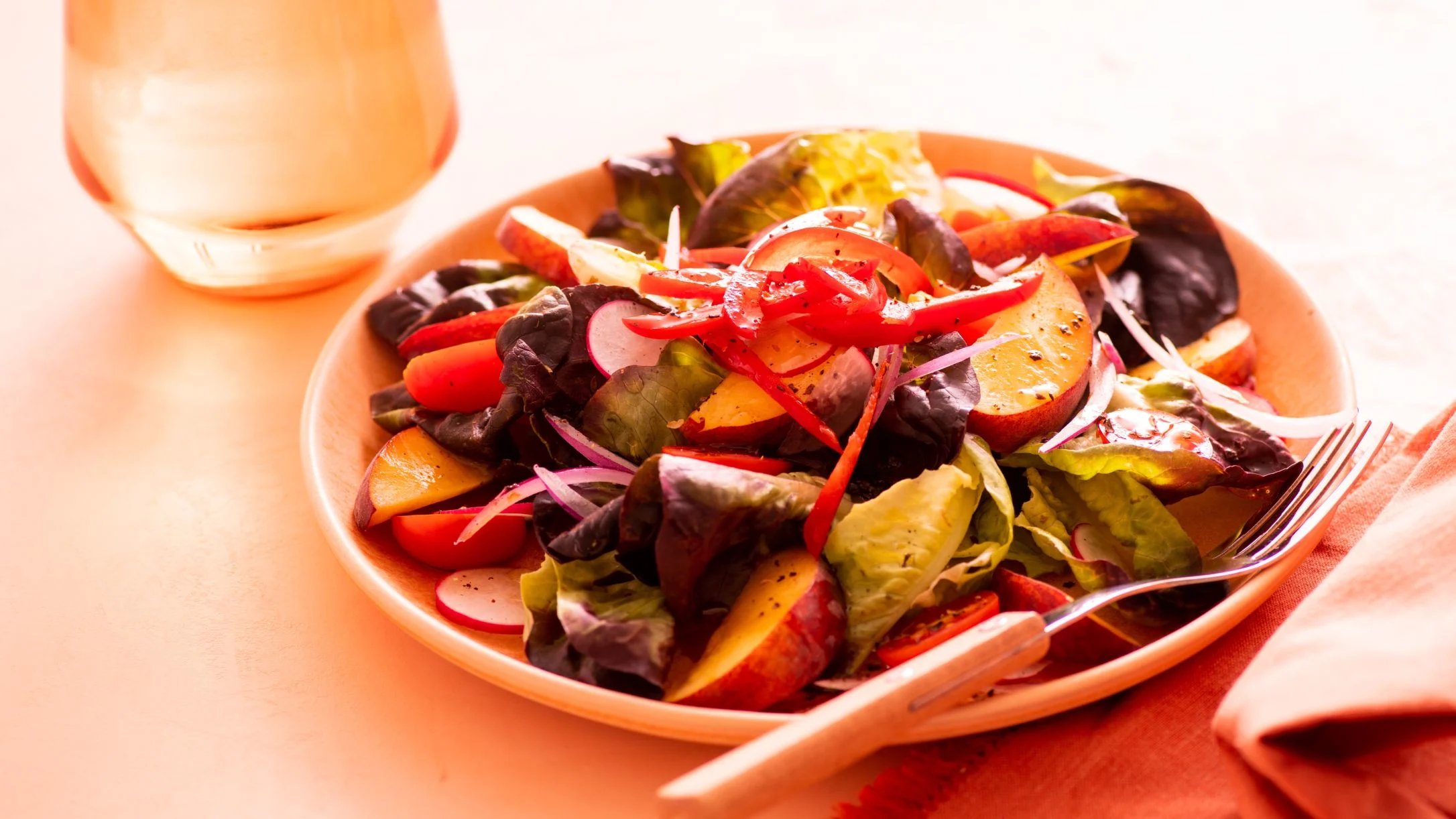 Fresh garden salad with mixed greens, sliced peaches, red peppers, and radishes on a peach-colored plate with a fork.