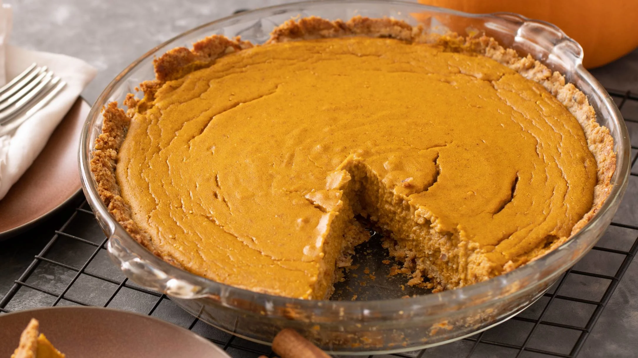 Freshly baked pumpkin pie in glass dish with slice removed, cooling on wire rack with fork nearby.