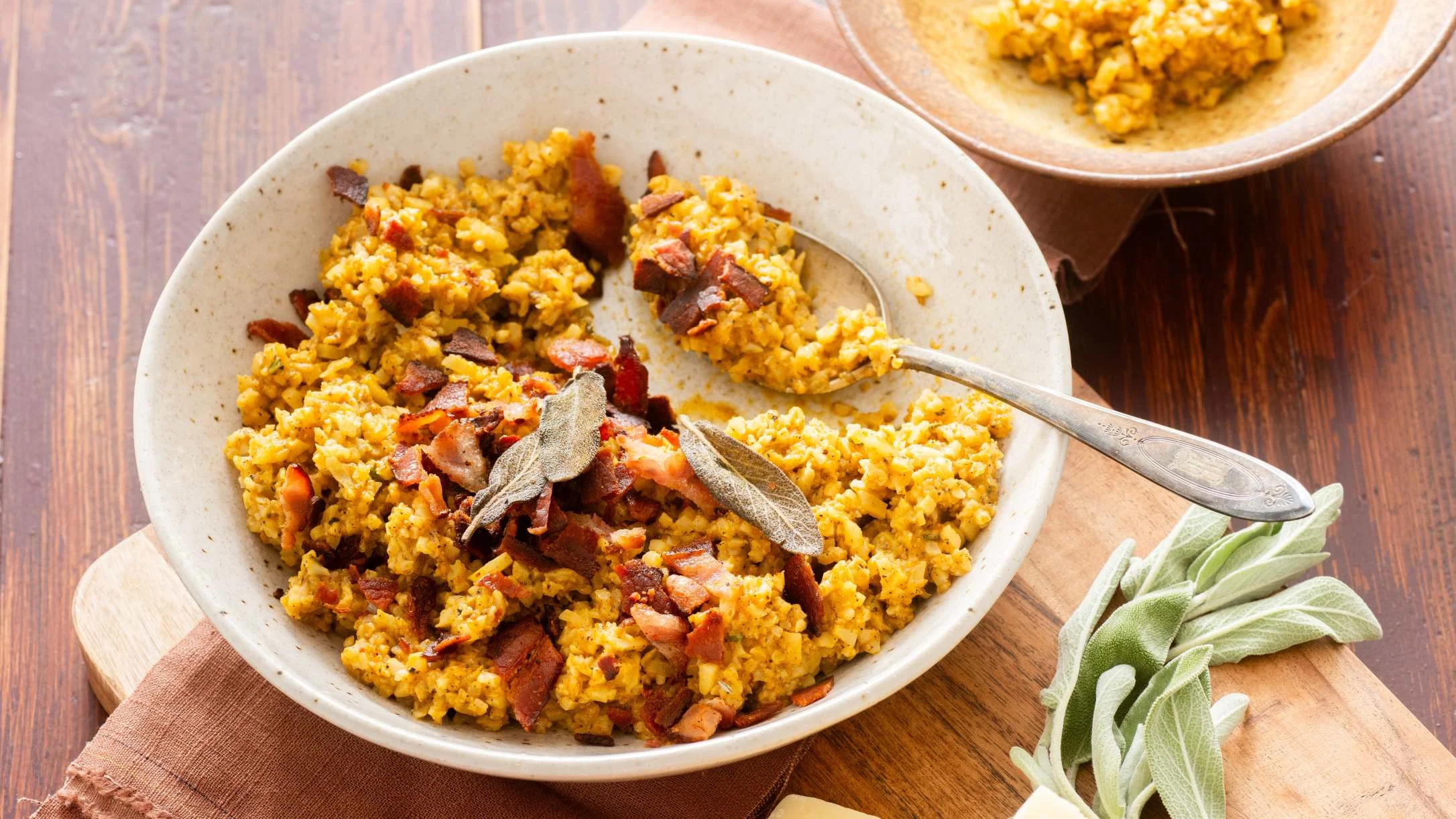 Yellow rice dish with bacon pieces and sage leaves in a ceramic bowl, served with a spoon on wooden board.