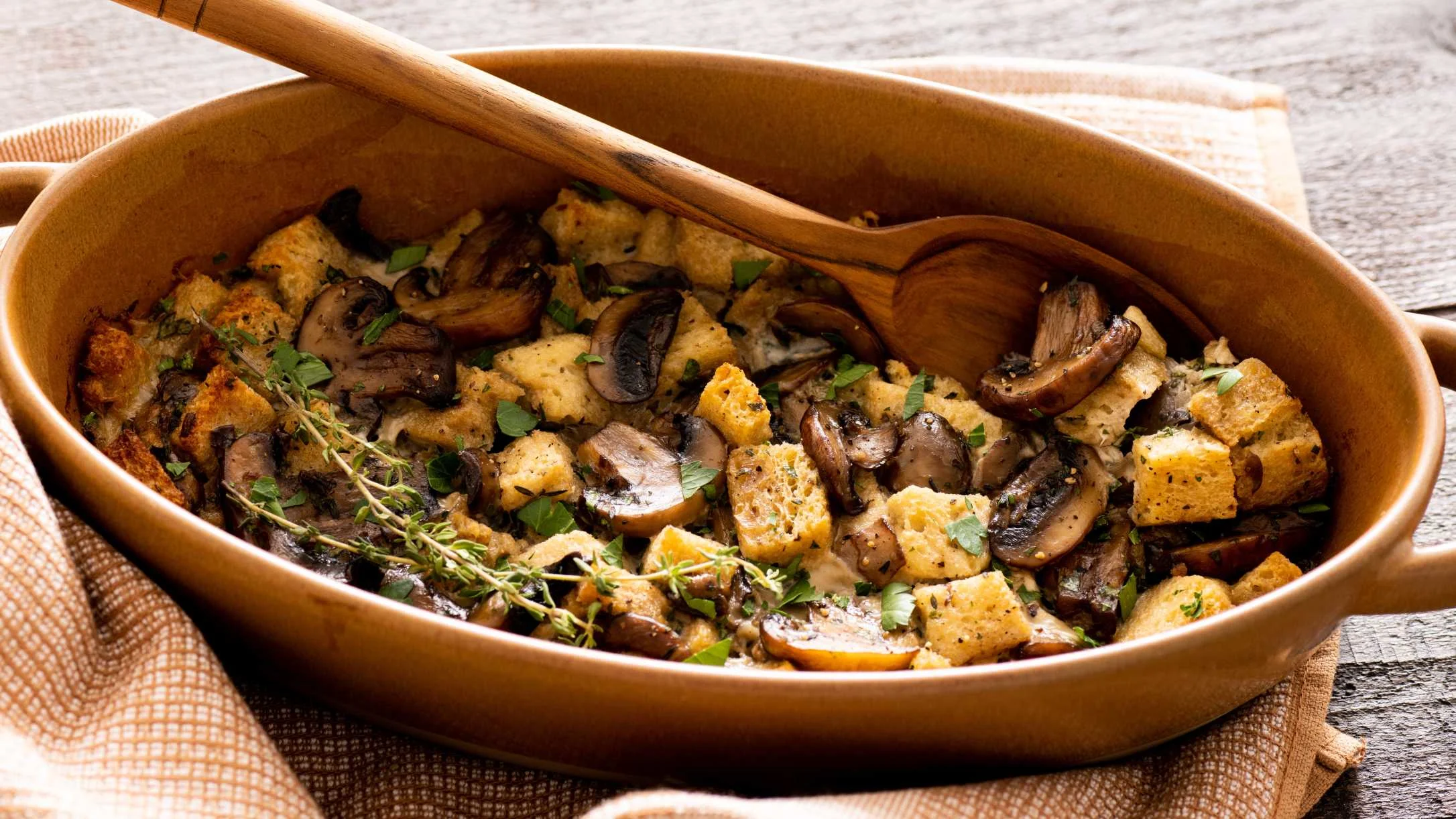 Mushroom bread stuffing in a brown ceramic dish with fresh herbs and wooden serving spoon.
