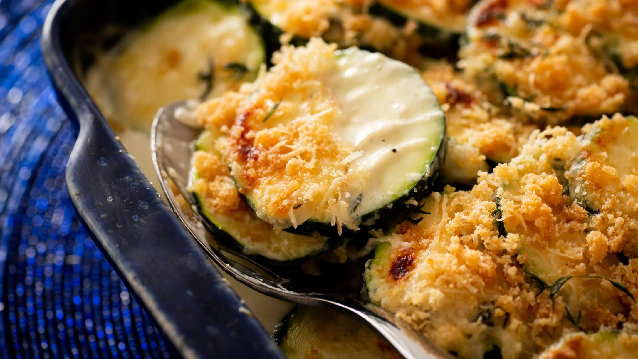 Baked zucchini gratin with golden topping and melted cheese in a blue ceramic dish.